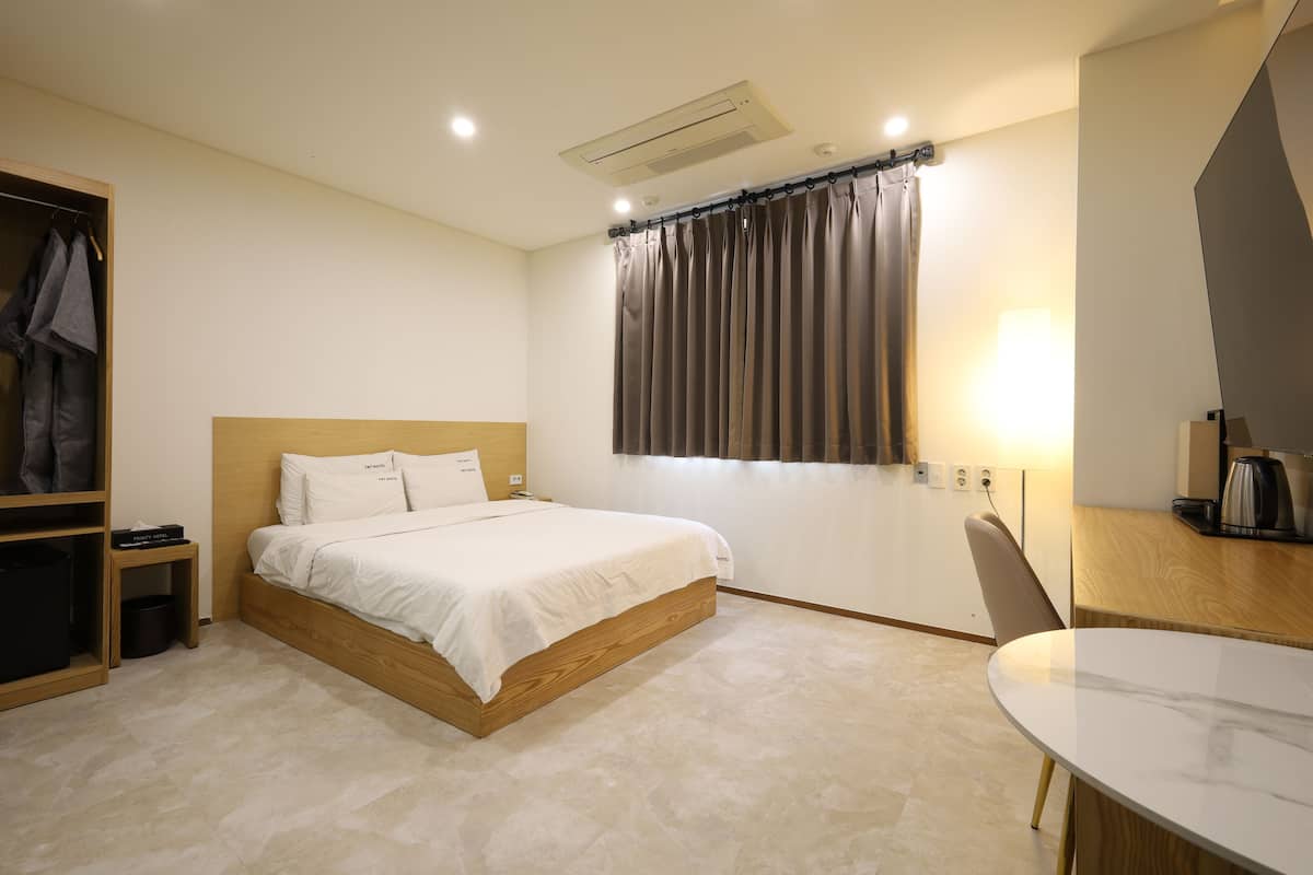 Une chambre d’hôtel comprenant un lit, une tête de lit en bois, une table de chevet, une chaise, une petite table, une fenêtre avec des rideaux et un climatiseur fixé au mur.