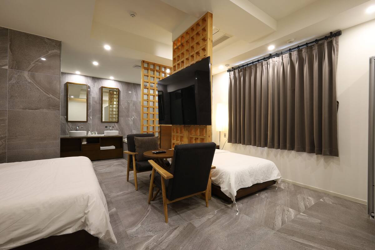 Une chambre d’hôtel avec deux lits, une télévision, une cloison en bois, un bureau avec une chaise et une salle de bain avec un miroir et un lavabo.