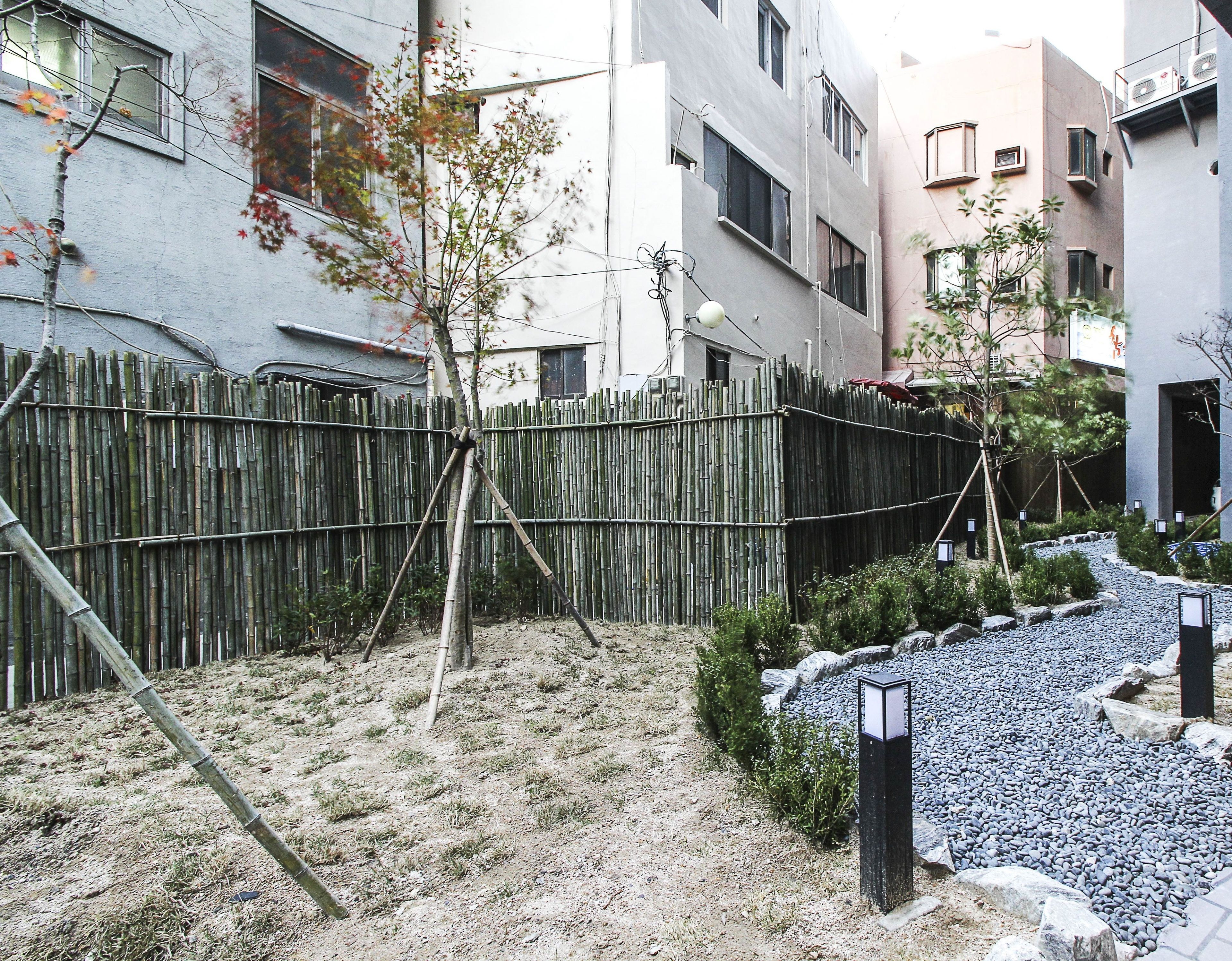 Zona residencial con valla de bambú, camino de grava y un pequeño jardín con plantas y rocas.