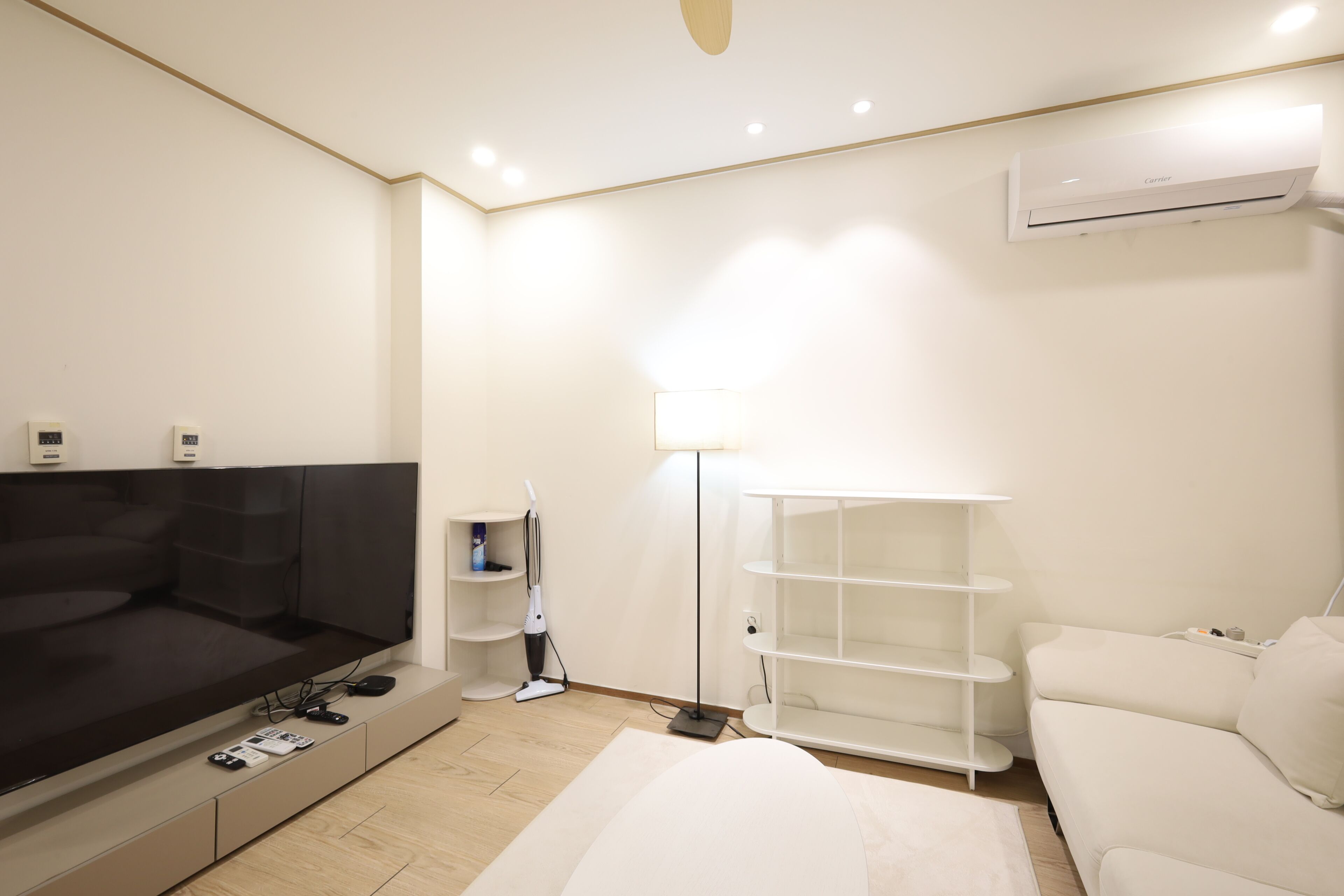 A modern hotel room with a flat-screen TV, a sofa, a standing lamp, a white air conditioning unit, and a white bookshelf.