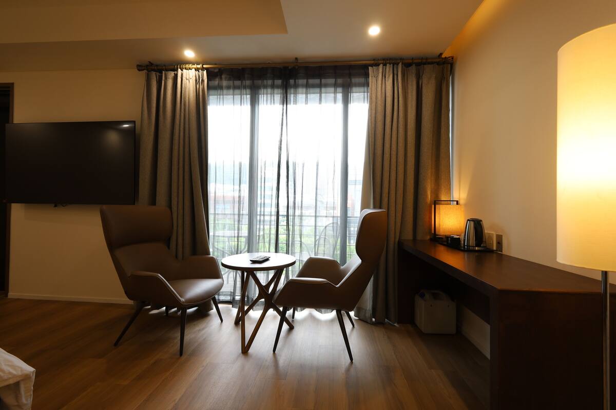 Une chambre d’hôtel moderne dotée d’une télévision à écran plat, de deux fauteuils, d’une petite table ronde, d’un bureau avec une lampe et offrant une vue sur l’extérieur grâce à de grandes fenêtres.