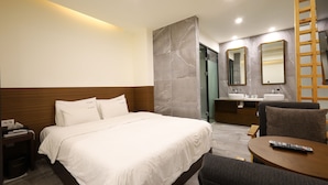 Una habitación de hotel moderna con una cama grande, una mesita de noche con lámpara, una pequeña mesa con control remoto y un baño con espejo y lavamanos.