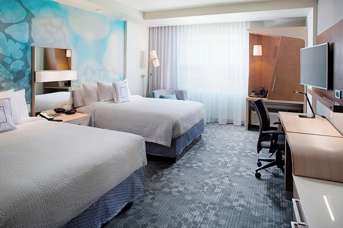 A hotel room with two beds, a desk, a TV, and a large window with curtains.