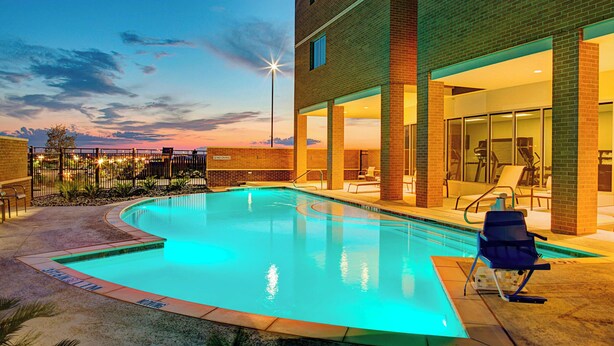 Una piscina de borde curvo, rodeada por un edificio de ladrillo con grandes ventanales y mobiliario exterior.
