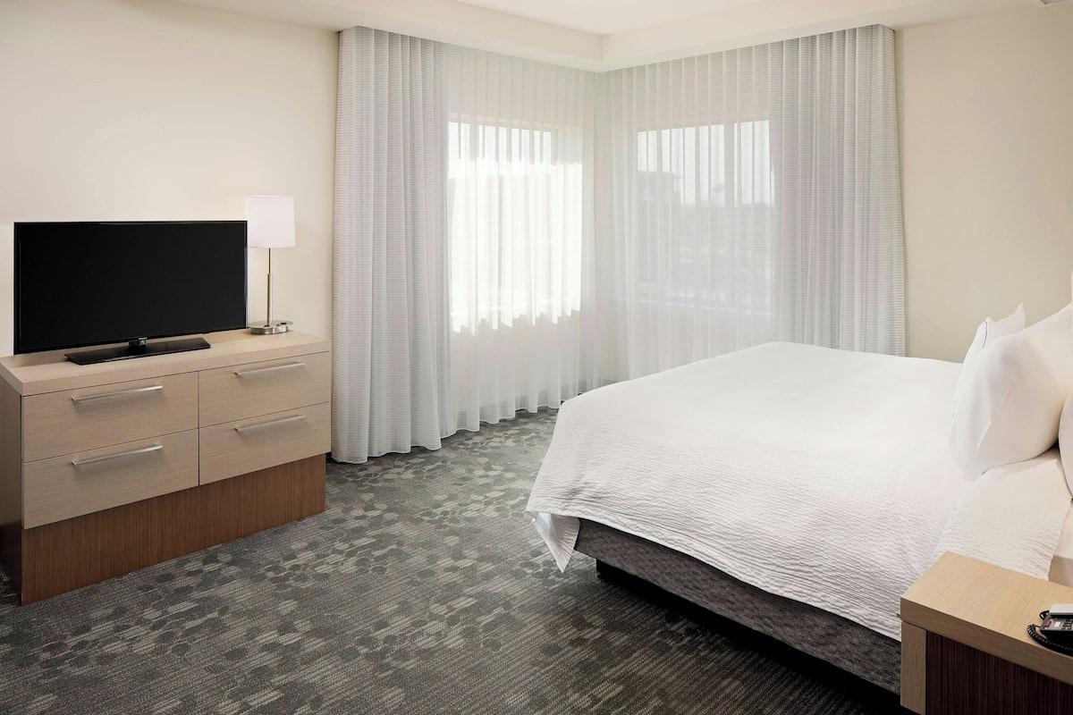 A hotel room with a bed, a TV on a wooden dresser, and a window with sheer curtains.