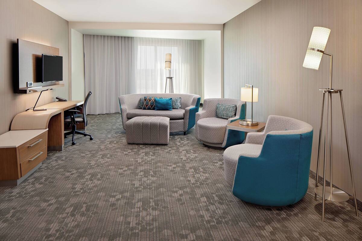 A hotel room with a flat-screen TV mounted on the wall, a desk with a chair, a sofa set, a standing lamp, and a large window with curtains.