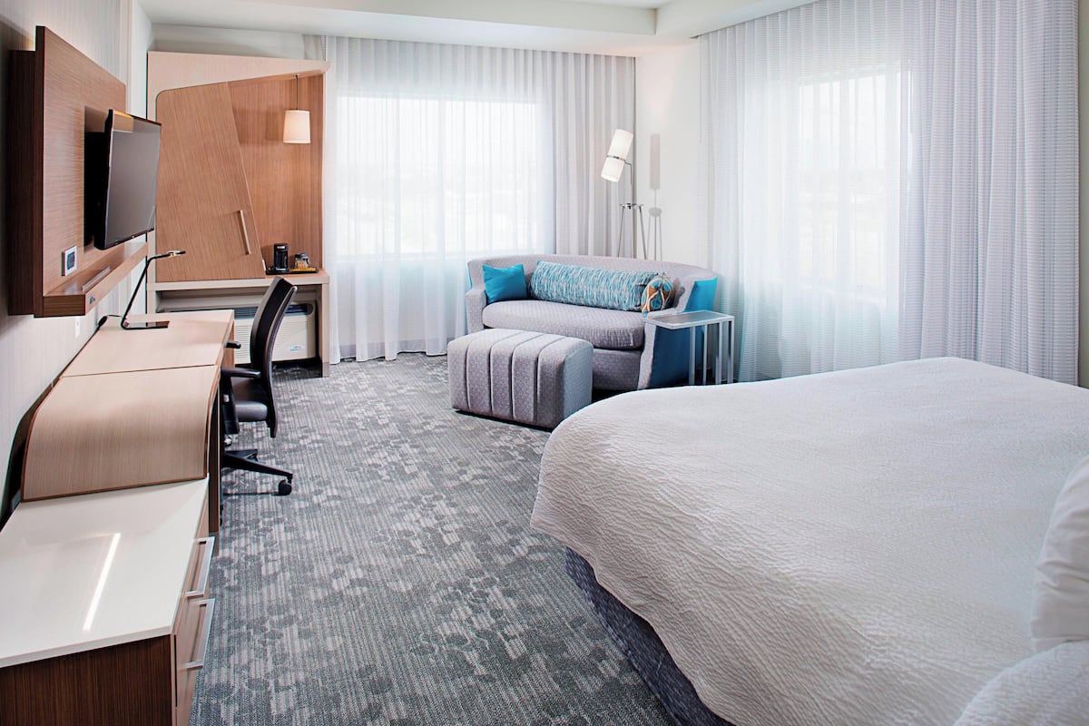 A hotel room with a bed, a desk with a chair, a TV, a sofa, and a lamp.