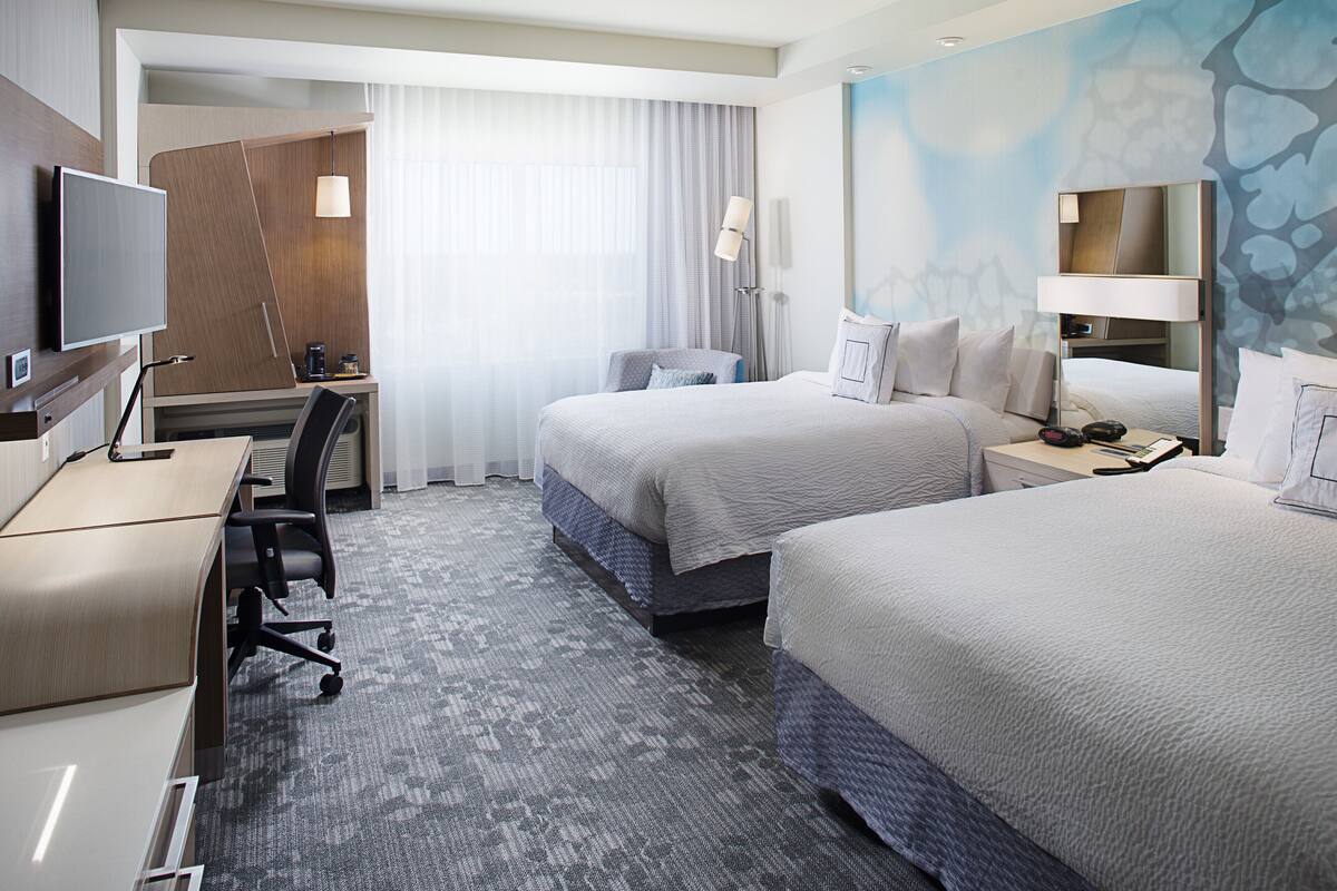 A hotel room with two beds, a desk, a chair, a TV, and a bathroom.