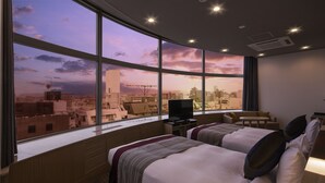 Habitación de hotel con dos camas, televisión y vistas a un paisaje urbano al atardecer.