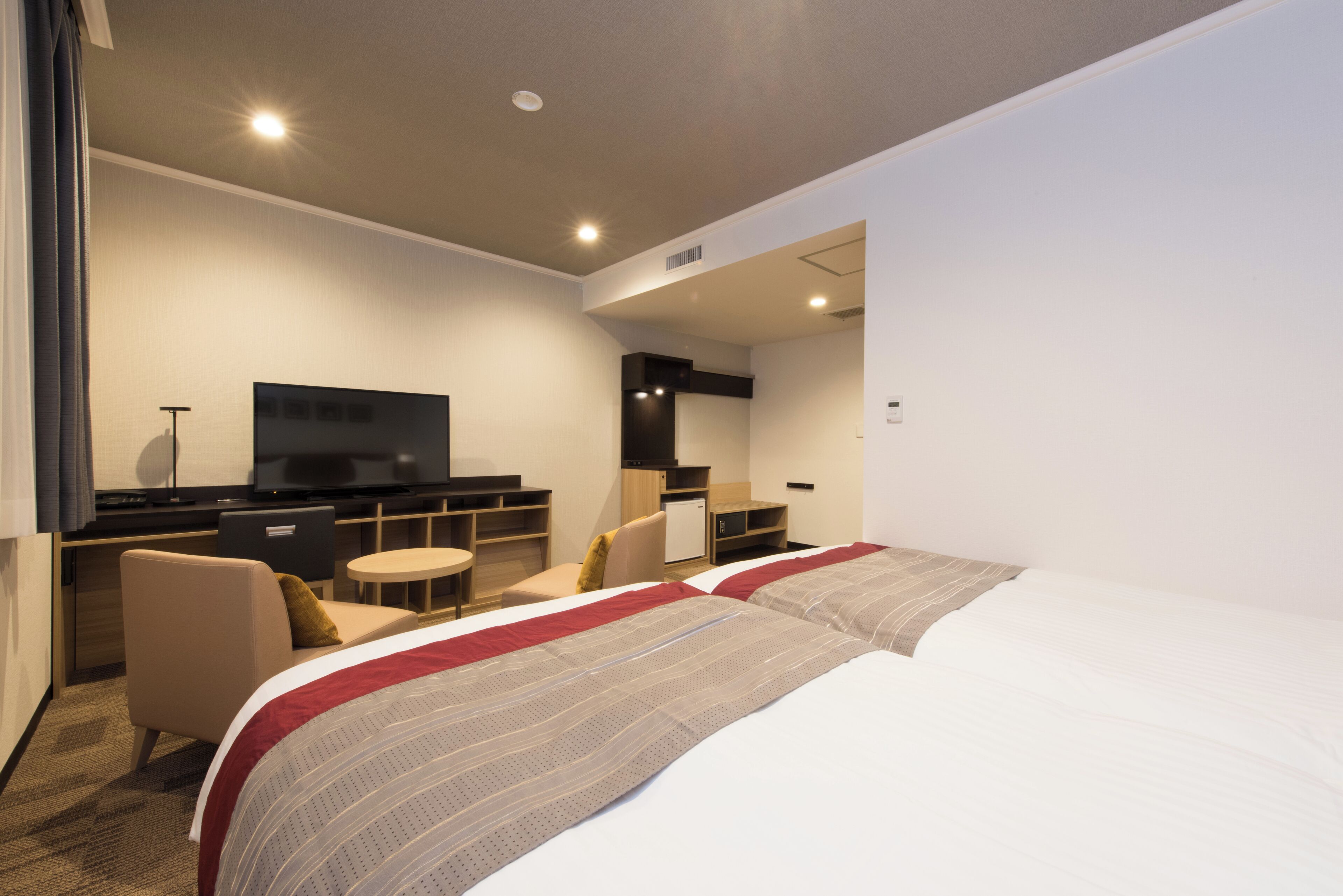 A modern hotel room with a large bed, a desk with a flat-screen TV, a small round table, and a comfortable armchair.
