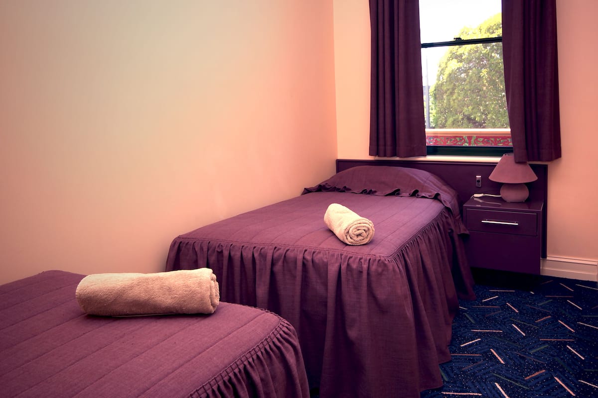 Una cama individual con colcha morada y una toalla enrollada, una mesita de noche con lámpara y una ventana con cortinas.