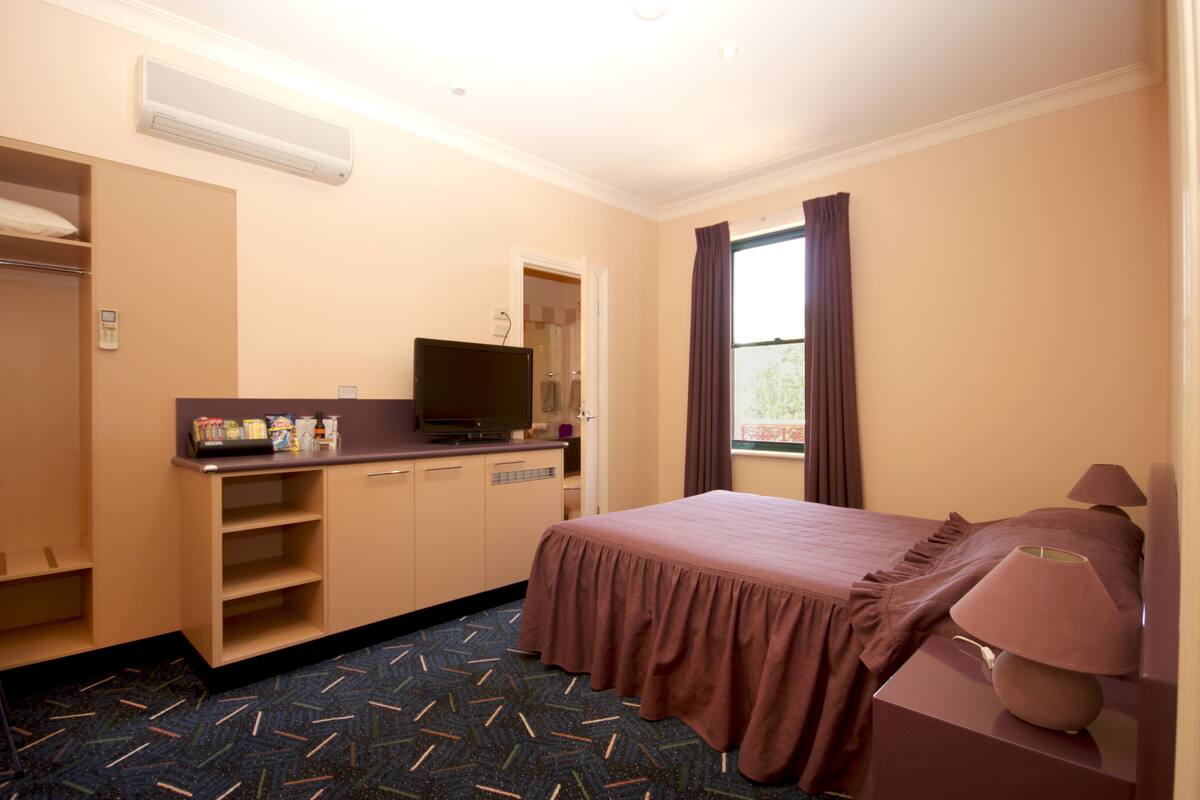 Habitación de hotel con cama, escritorio con televisor, ventana con cortinas y unidad de aire acondicionado.