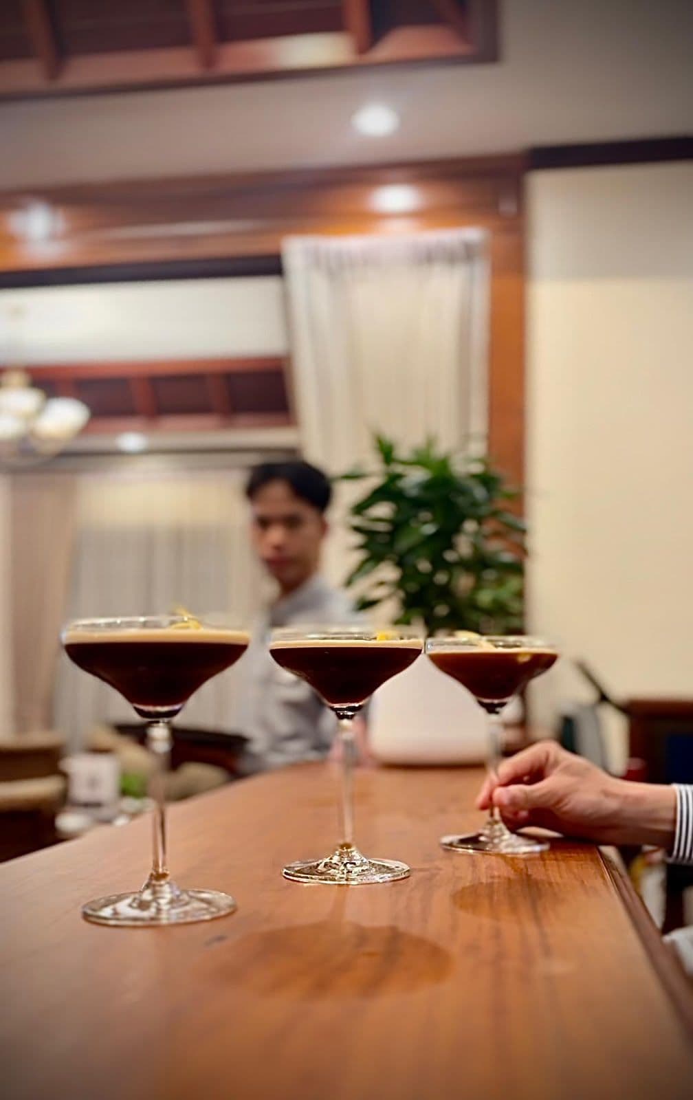 A bar area with three martini glasses on a wooden counter, a person’s hand reaching for one, and a blurred figure in the background.