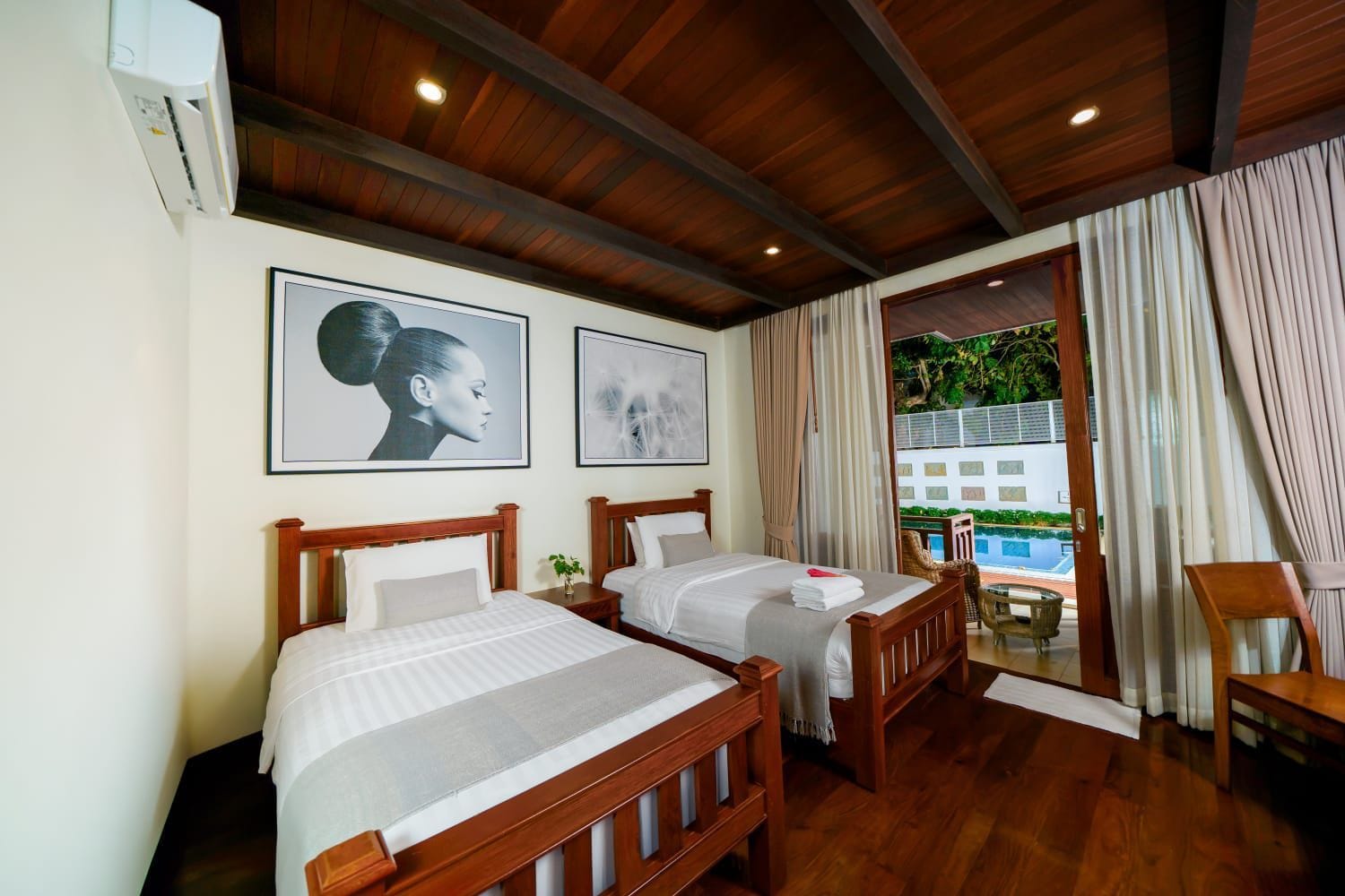 Habitación de hotel con dos camas, cabeceros de madera, un ventanal con cortinas y vista a una piscina.
