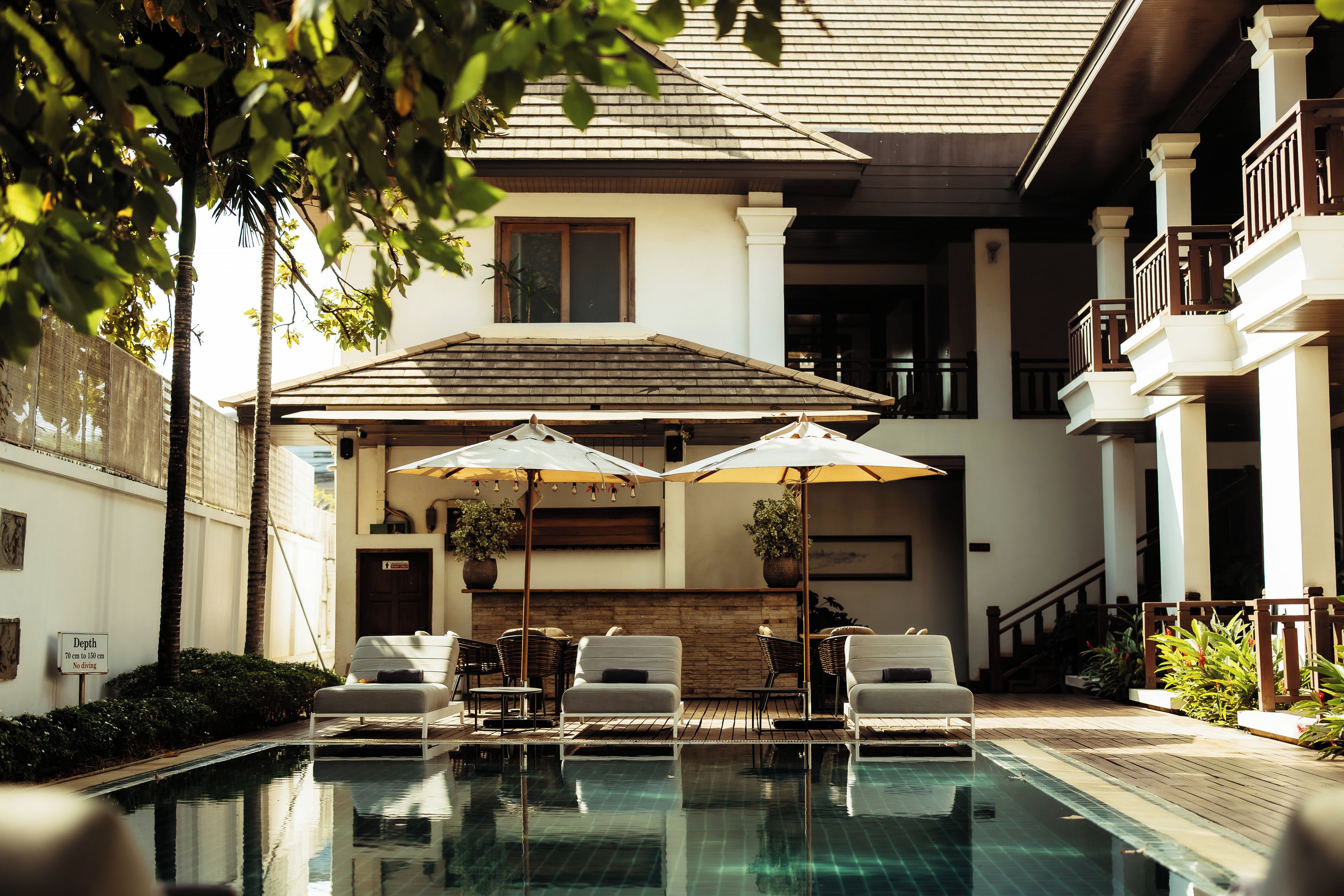 Un espace piscine avec des chaises longues et des parasols, une cheminée en pierre et un bâtiment à deux étages doté d’un balcon.
