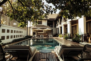 An outdoor swimming pool with lounge chairs and umbrellas, surrounded by a building with a balcony and a wall of artwork. - Athena Hotel (Pakse)