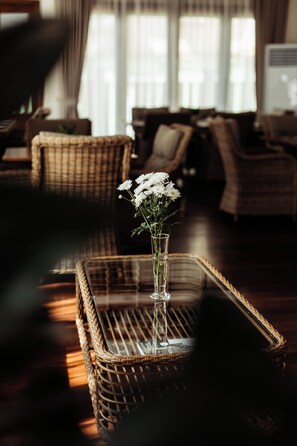 A wicker chair with a vase of flowers in a room with a large window and a sofa. - Athena Hotel (Pakse)
