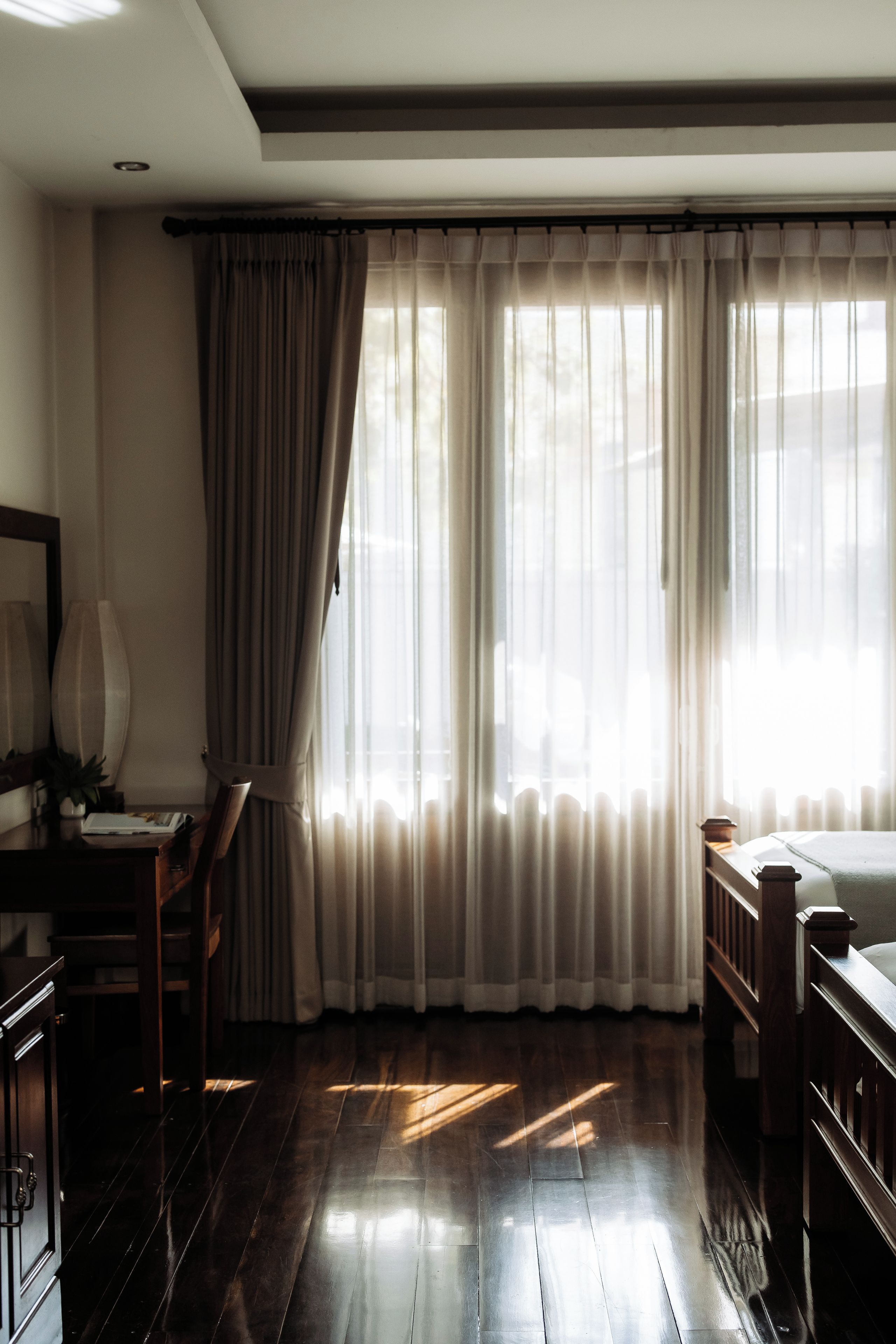 A room with a wooden floor, a desk with a chair, a bed, and a window with sheer curtains.