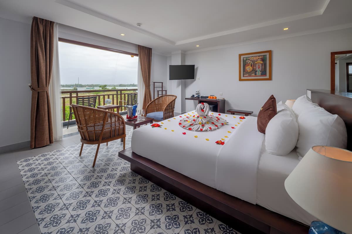 Quarto de hotel com uma cama grande, uma varanda com cadeiras de vime, uma televisão e um quadro na parede.