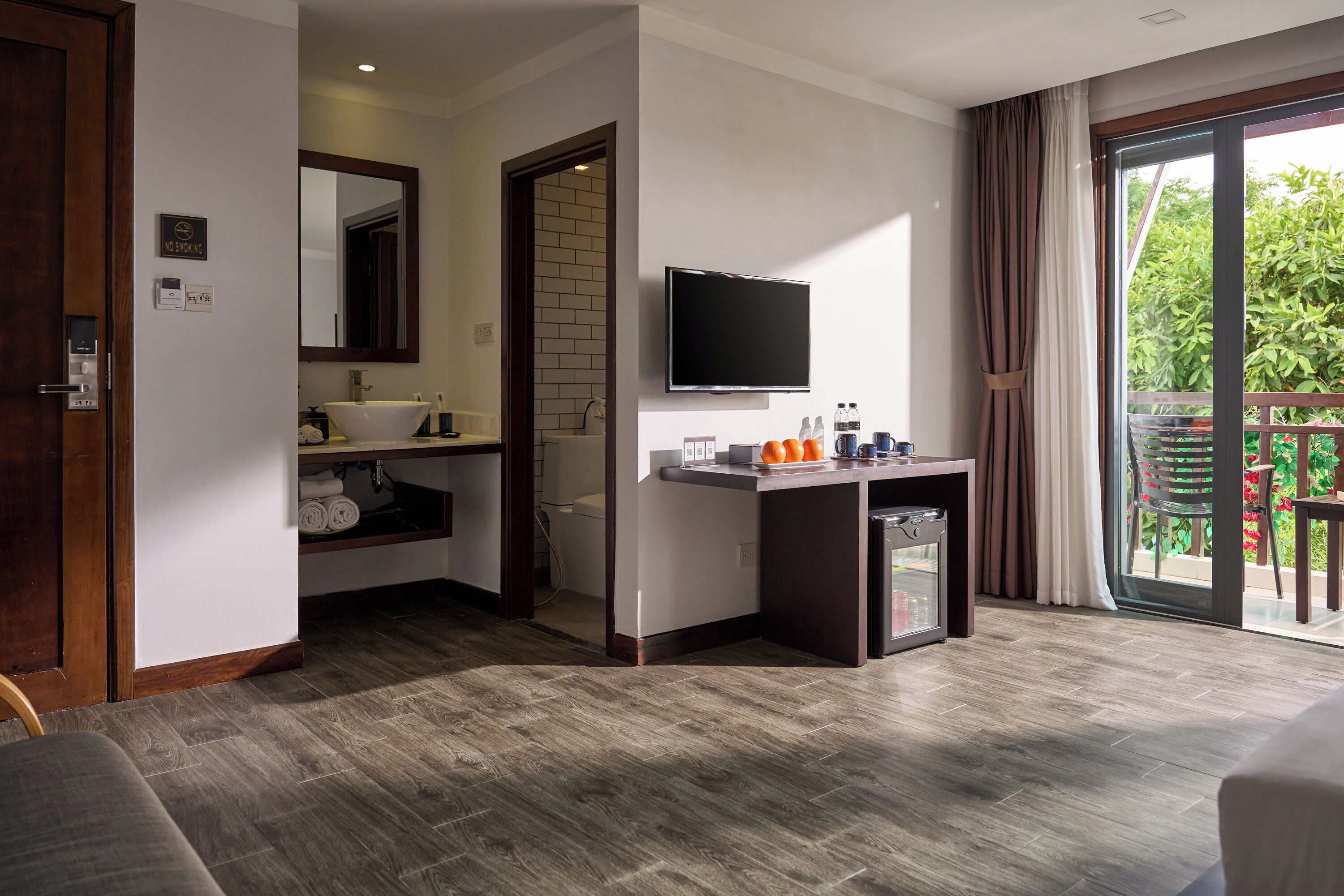 A modern hotel room with a wooden floor, a large mirror, a flat-screen TV, a small desk with a sink, and a balcony with a view of greenery.