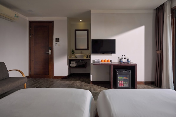 A hotel room with two beds, a TV, a small desk with a mirror, a shelf with toiletries, and a window with curtains. - Hoi An Odyssey Hotel (Da Nang)
