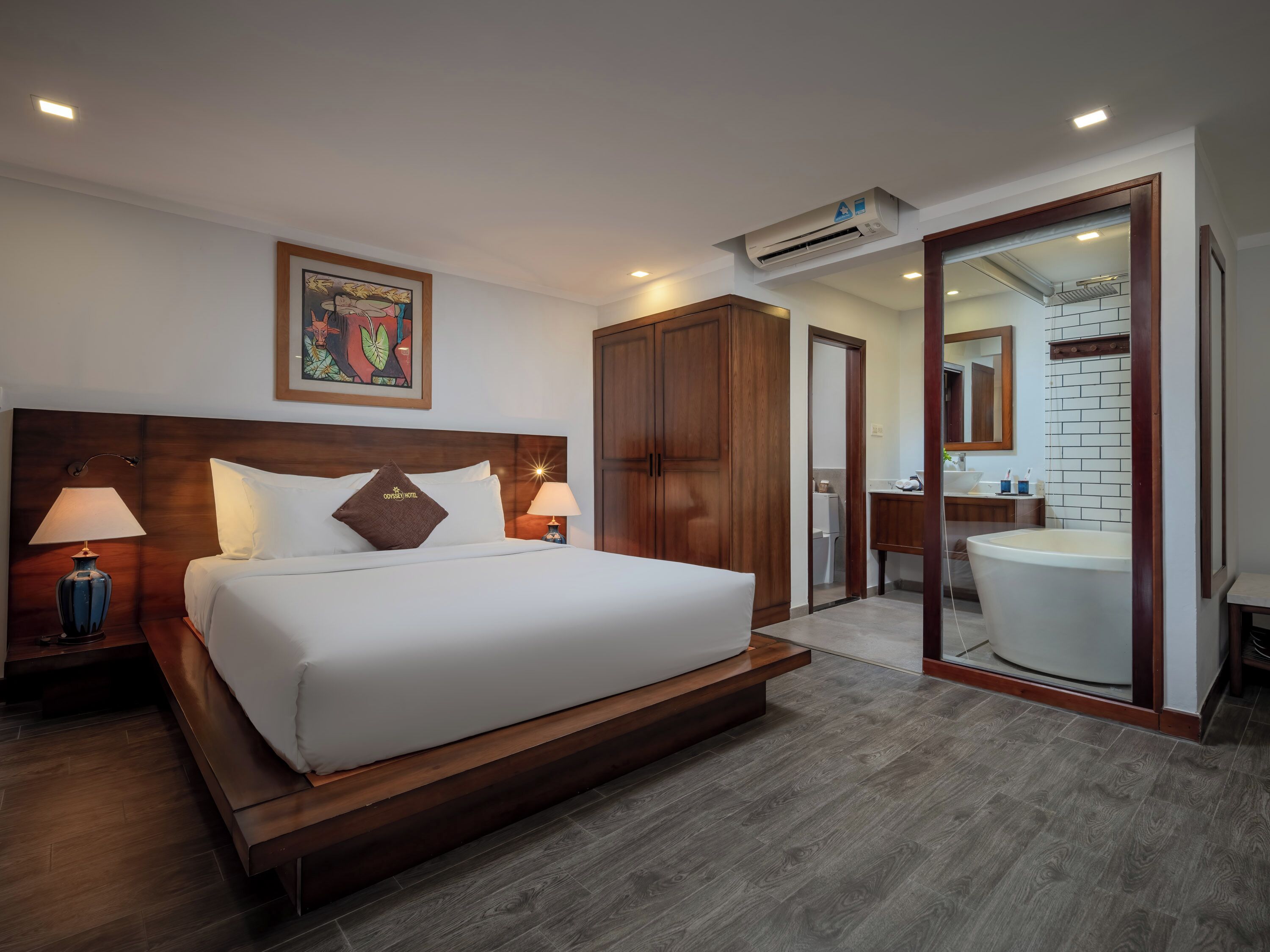 A hotel room with a large bed, wooden headboard, two bedside lamps, a nightstand, a painting on the wall, a wardrobe, an air conditioning unit, a bathroom with a sink, mirror, and a large bathtub.