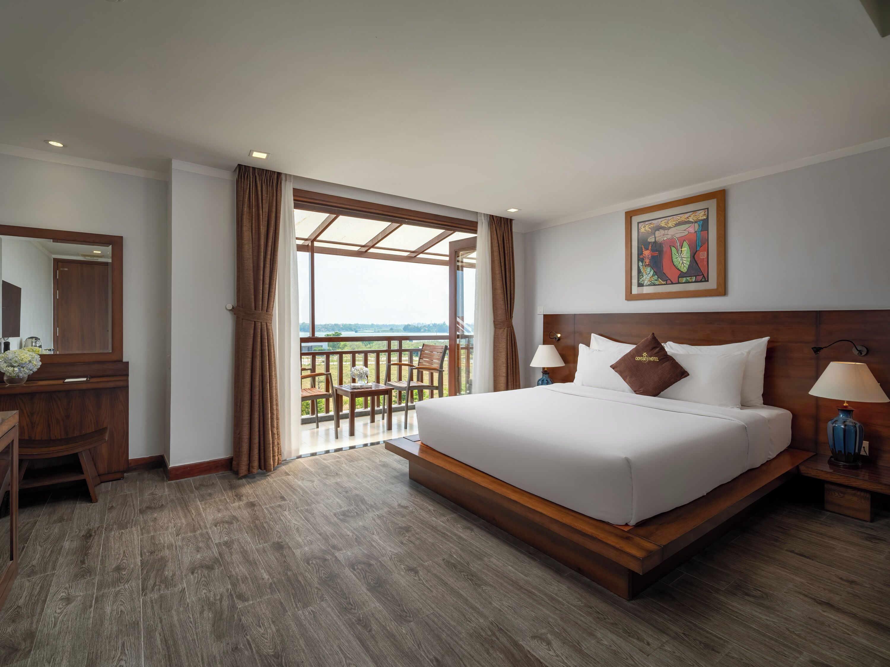 A hotel room with a large bed, a wooden desk, a mirror, and a balcony with a view.