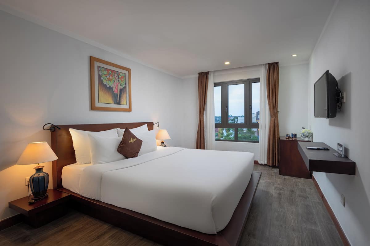 Quarto de hotel com uma cama grande, uma escrivaninha, uma televisão e uma janela com cortinas.
