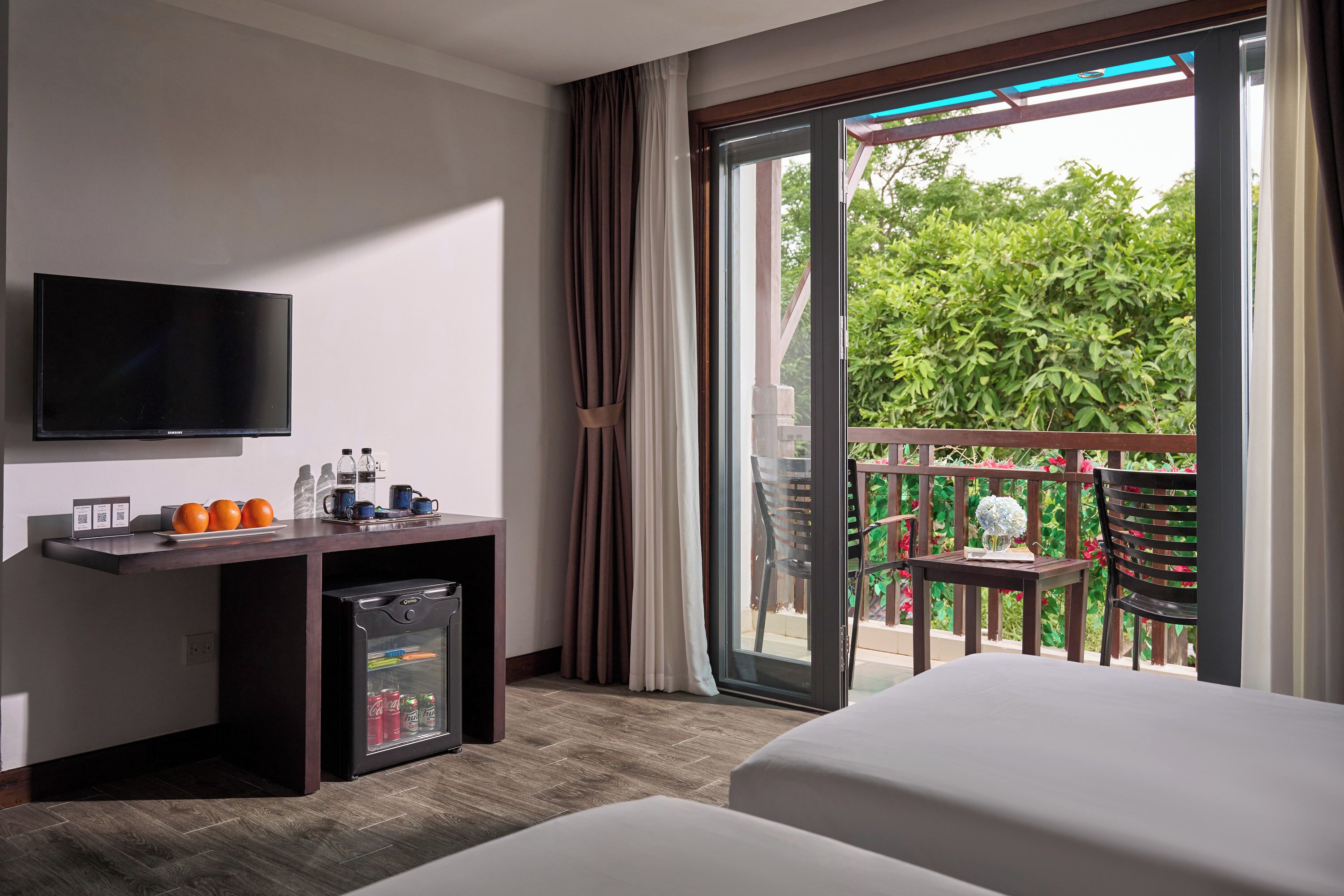 Une chambre d’hôtel comprenant un lit, un bureau avec une télévision, un mini-réfrigérateur et un balcon donnant sur un espace verdoyant.