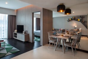 Una habitación de hotel moderna con zona de comedor, televisión y un gran ventanal con cortinas.