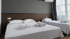 Habitación de hotel con dos camas, pared de textura gris, paneles de madera, unidad de aire acondicionado y ventana con cortinas translúcidas.