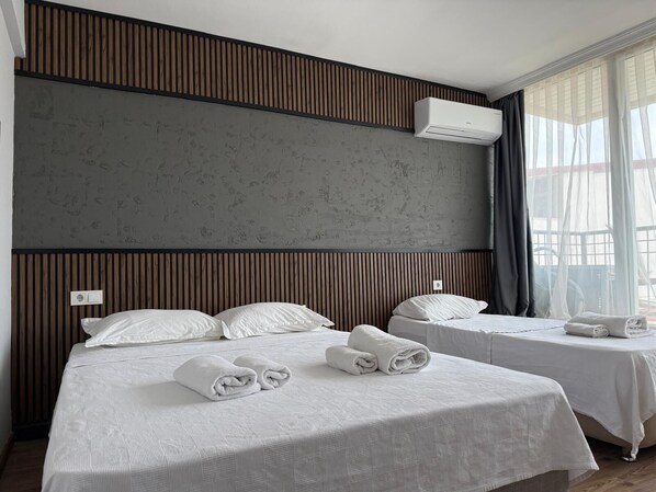 A hotel room with two beds, a grey textured wall, wooden paneling, an air conditioning unit, and a window with sheer curtains.