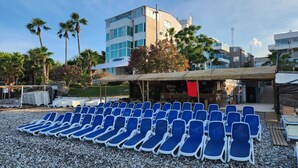 Un hotel frente al mar con filas de sillas azules para tomar el sol, un área de estar cubierta al aire libre y edificios modernos al fondo.