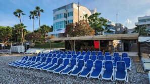 A beachside hotel with rows of blue lounge chairs, a covered outdoor seating area, and modern buildings in the background. - Romeo Beach Hotel (Kemer)