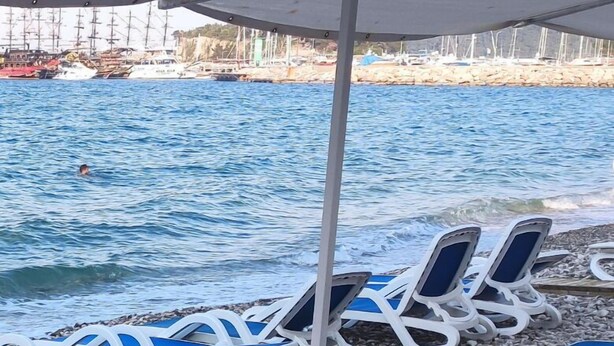 Una fila de tumbonas con cojines azules y marcos blancos, dispuestas en una playa de piedrecitas con vista despejada al mar y barcos al fondo.