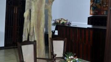 Una habitación con una estatua de madera de una figura, dos sillas de madera, una mesita con un arreglo floral y un ventilador de techo.