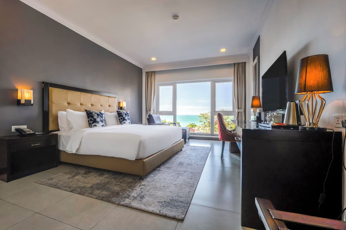 A modern hotel bedroom with a large bed, a TV, a desk, and a view of the sea.