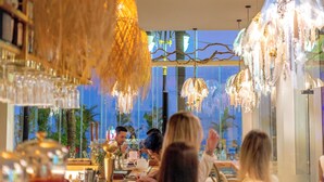Een eetruimte met meerdere hangende kroonluchters, een groot raam met uitzicht op palmbomen, en een bar met flessen op de planken.