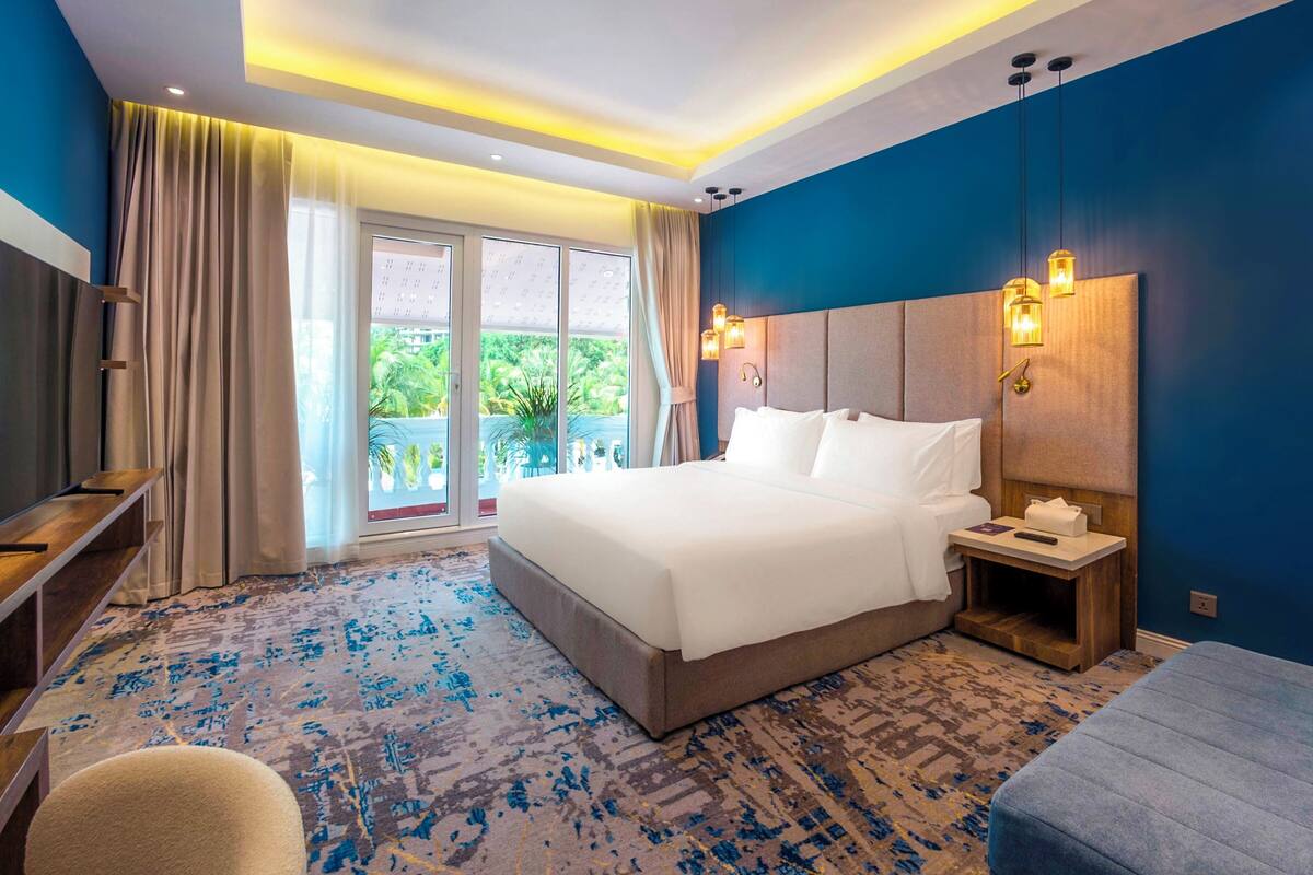 Ein Hotelzimmer mit einem großen Bett, einem Nachttisch, einem Fernseher und Blick auf Grünfläche.