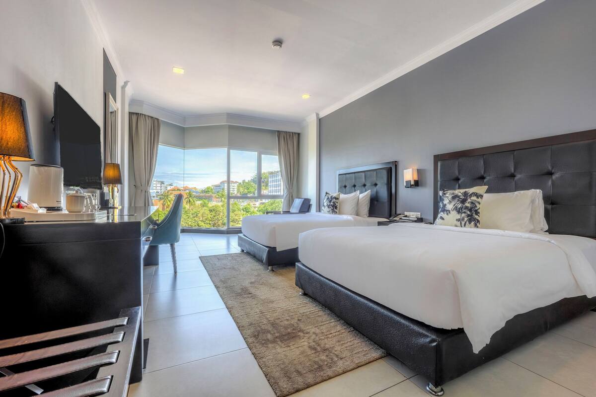 A modern hotel room with two beds, a flat-screen TV, a desk with a lamp, and a view of the city.
