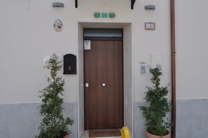 Una puerta de madera con felpudo, dos plantas en macetas y un letrero con número que indica la entrada.