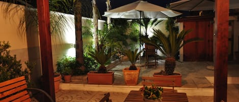 Un patio al aire libre con bancos y mesas de madera, rodeado de plantas en macetas y sombrillas.