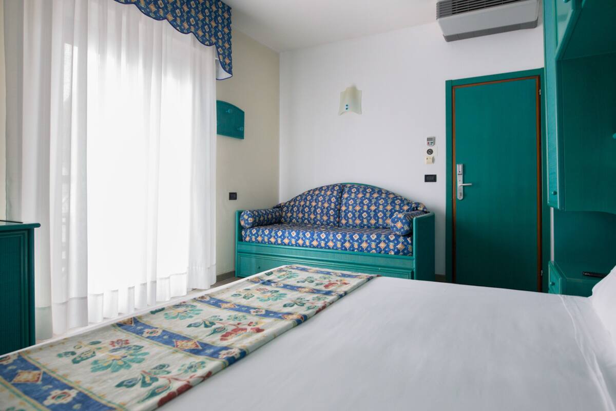 A room with a bed, a patterned bench, a teal door, and a window with sheer curtains.