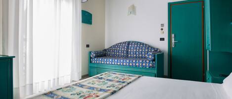 Una stanza con un letto, una panca decorata, una porta color petrolio e una finestra con tende leggere.