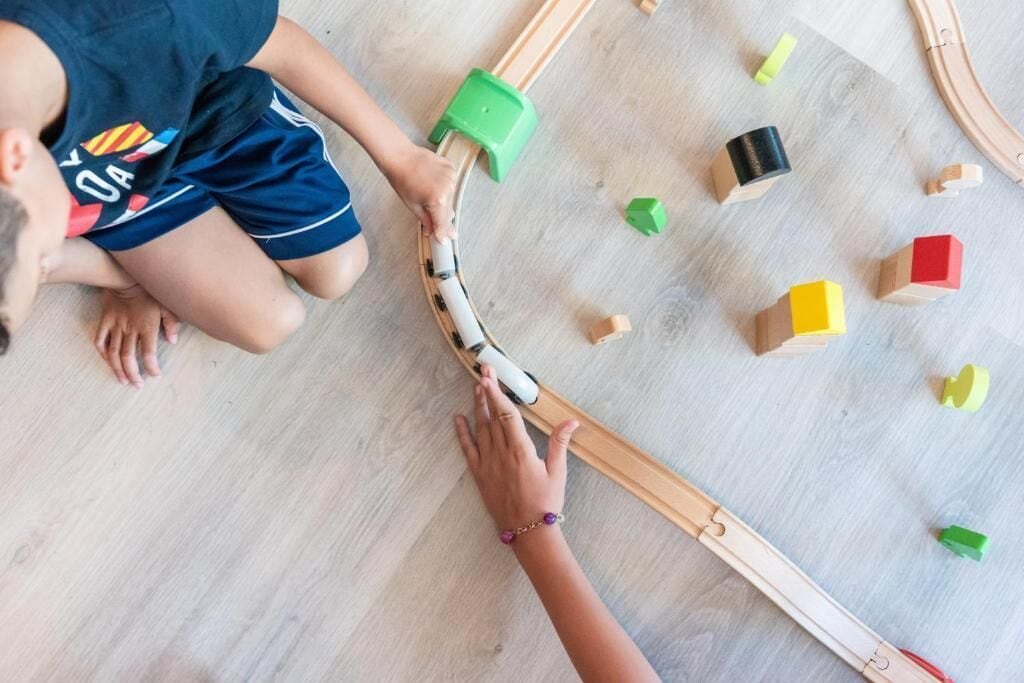 A child playing with wooden train tracks and blocks on a wooden floor.