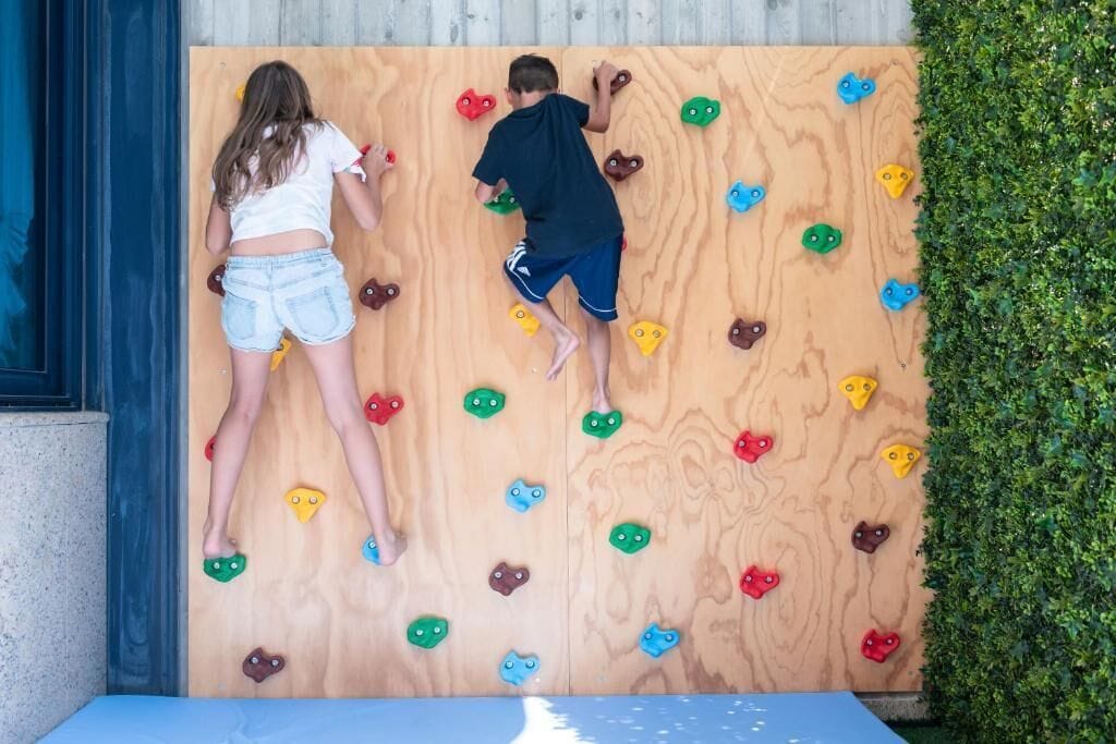 Two children climbing a wooden wall with colorful holds.