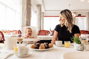 Un comedor con una mesa dispuesta para dos personas, que incluye una trona para un niño, diferentes platos y bebidas.