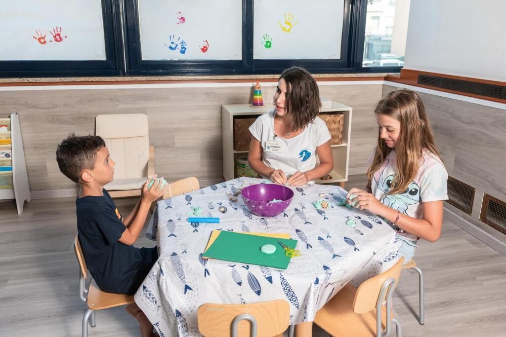A room with a table where three individuals are engaged in an activity, a window with artwork, and a shelf with items.