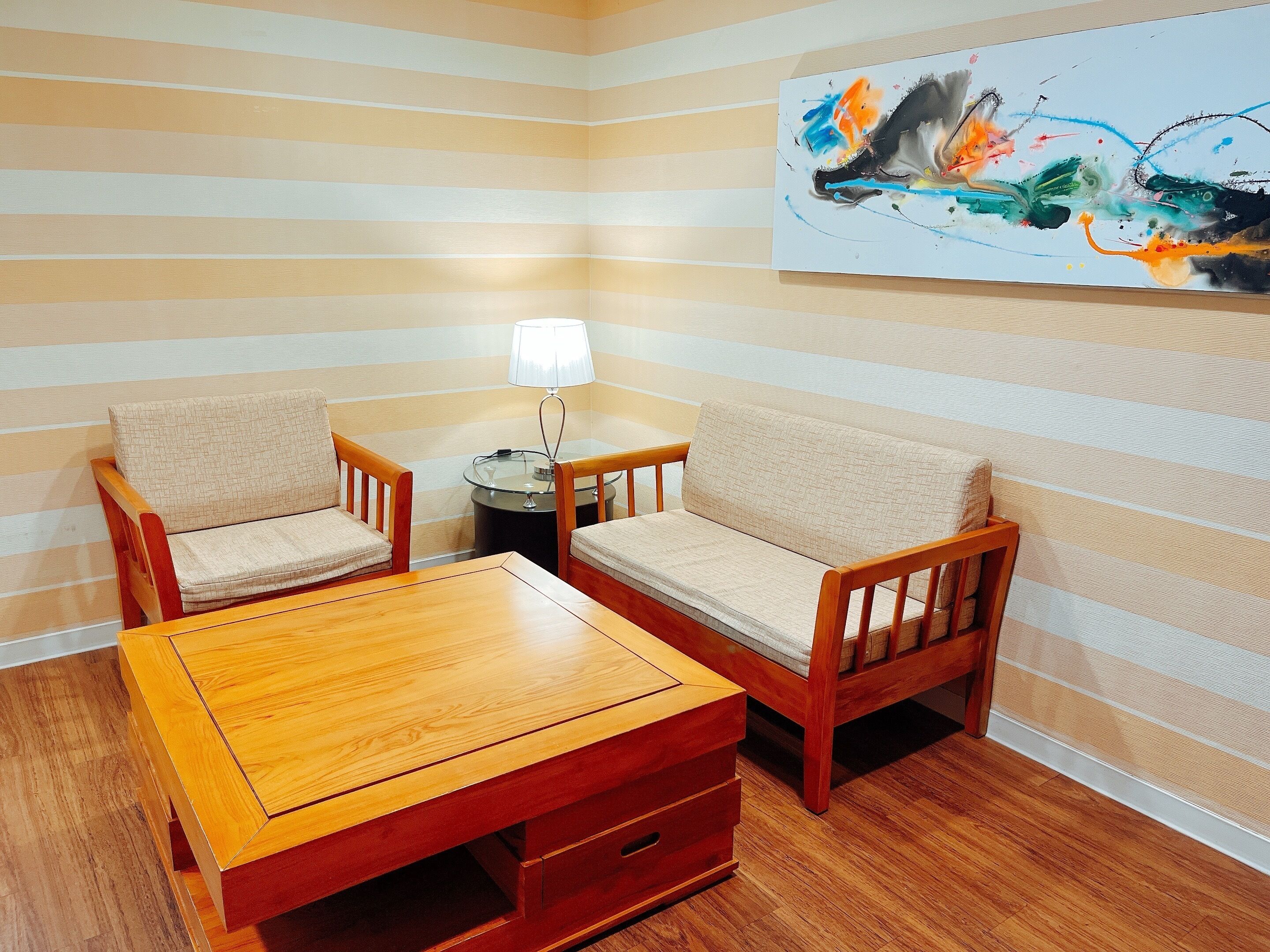 A corner room with striped walls, a wooden coffee table, two armchairs, a small round table with a lamp, and a colorful abstract painting on the wall.