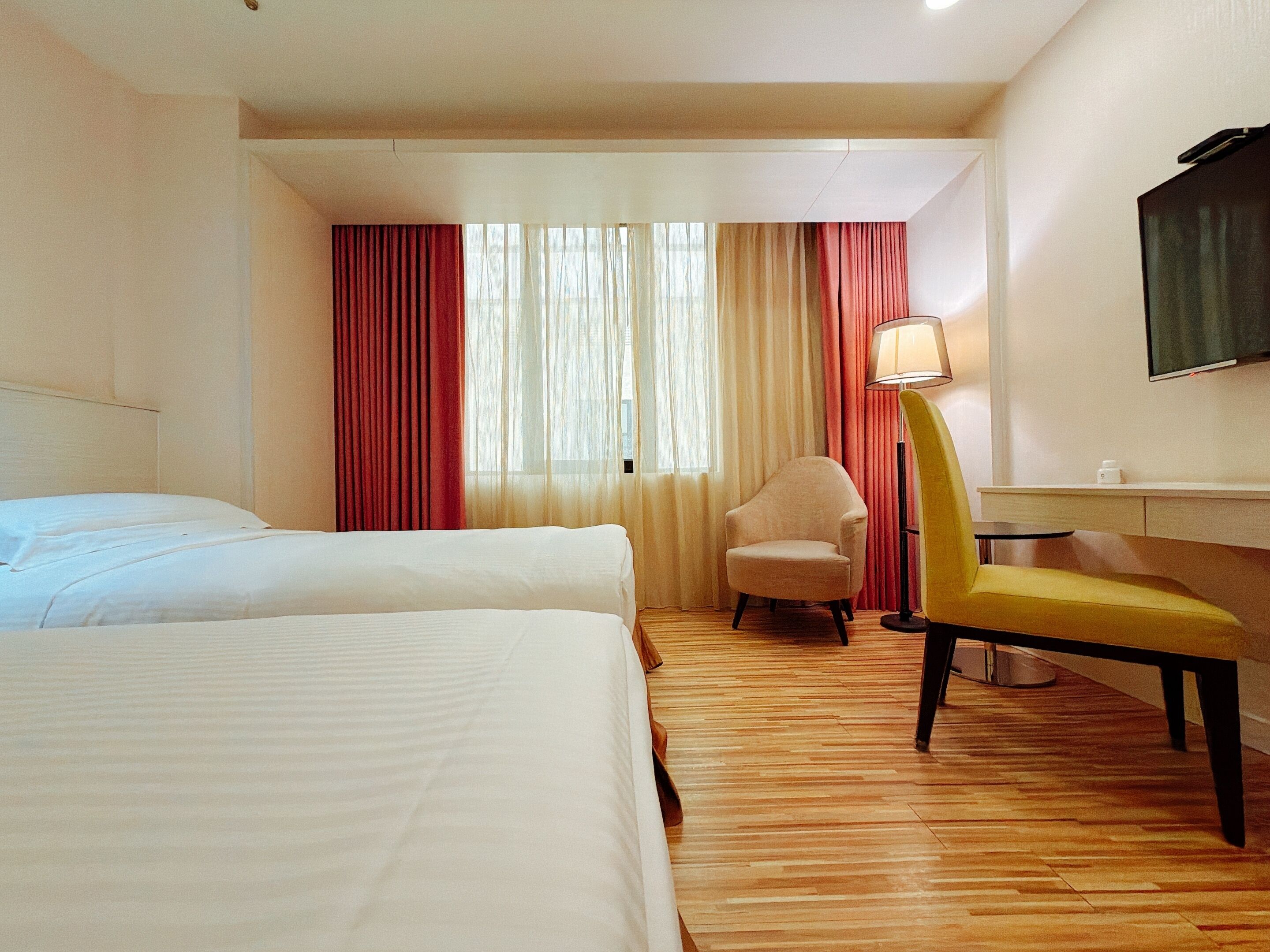 A hotel room with two beds, a desk, a chair, a lamp, and a TV on the wall.